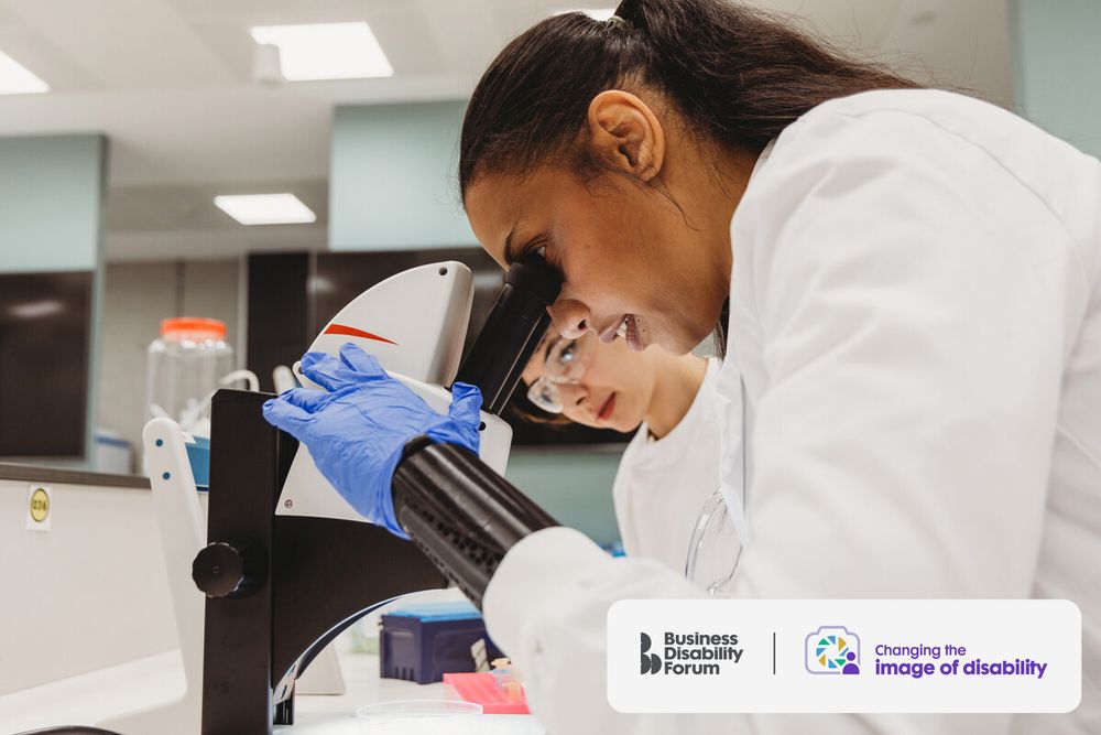 Scientist wearing a prosthetic hand on the left side looks into a microscope in a lab, with a colleague next to her. The Business Disability Forum 'Changing the image of disability' logo is in the lower right corner of the image.
