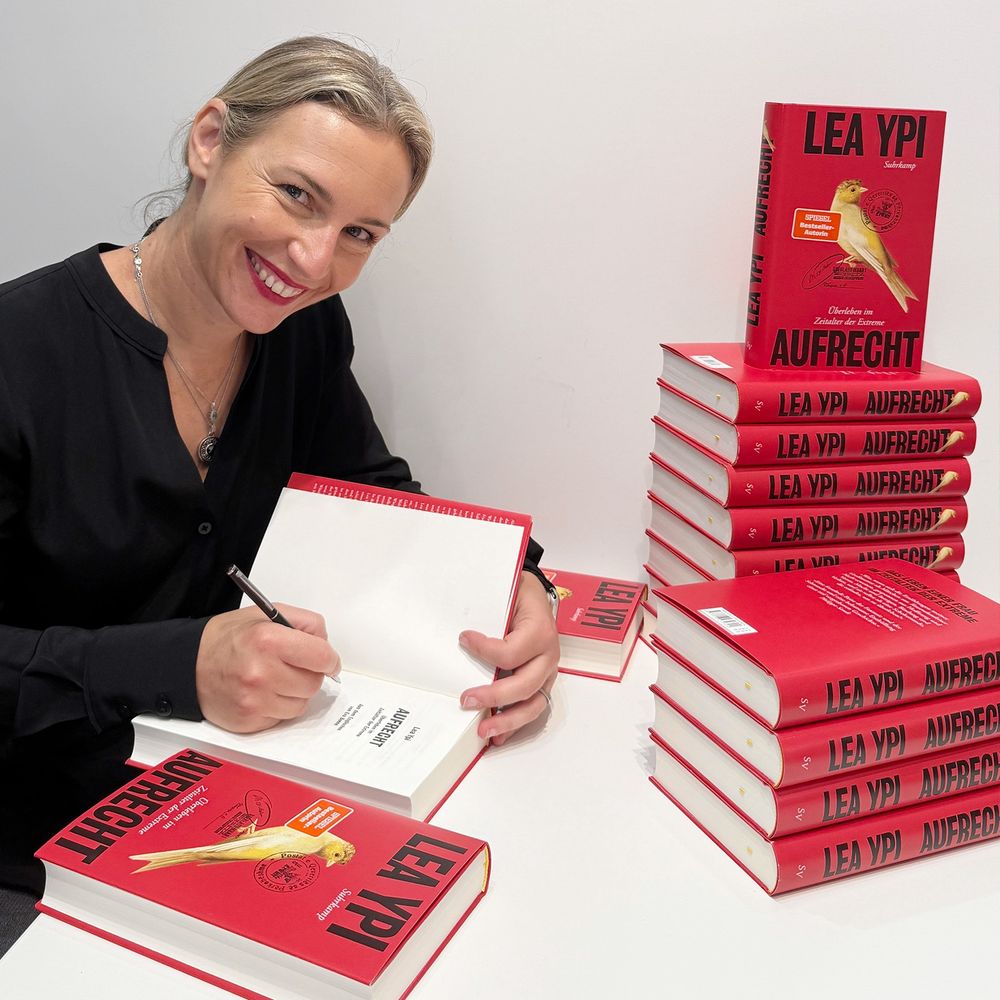 Lea Ypi sitzt an einem Tisch, auf dem mehrere Stapel ihres Buchs »Aufrecht« liegen und lächelt in die Kamera während sie eine Ausgabe signiert.