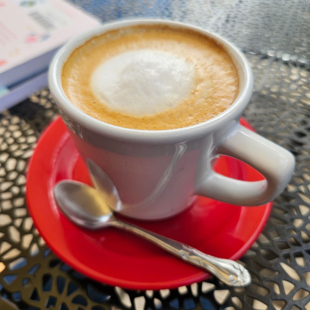 A small white coffee cup with coffee colored foam and around white circle of foam in the middle, sitting on an orange coffee plate, with a small silver colored shiny spoon resting on the plate, which is on a black perforated metal garden table, with a couple of books nearby.