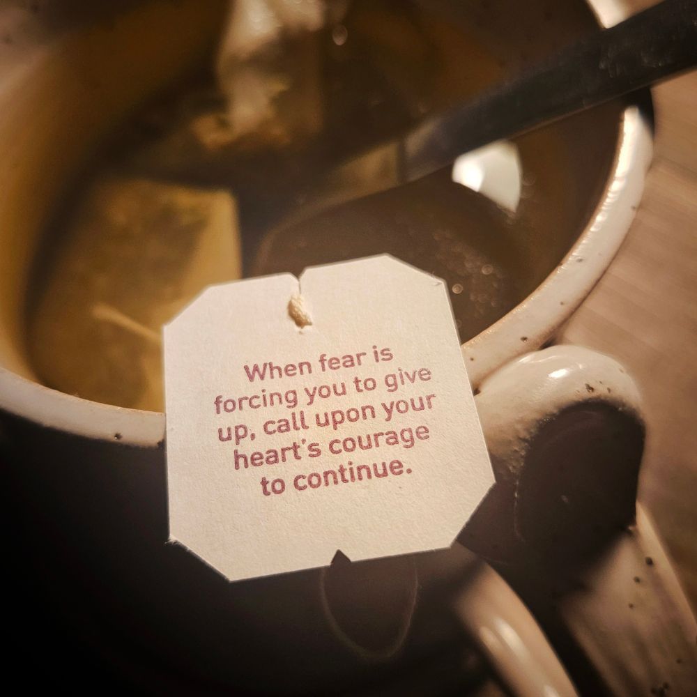 A close-up at night with the light of a desk lamp out of the frame, illuminating a hand crafted off-white ceramic mug with a teabag inside with yellow tinged tea and the tea tag attached to a string balanced on the edge that reads: When fear is forcing you to give up,  call upon your heart's courage to continue. 