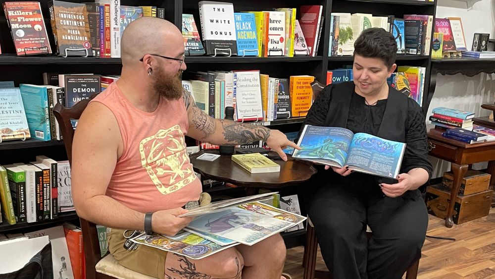 Matthew Spencer and Ava Kelly are sitting next to a small round table with the Alia Terra book and original illustrations, while Matt points at the open book.
