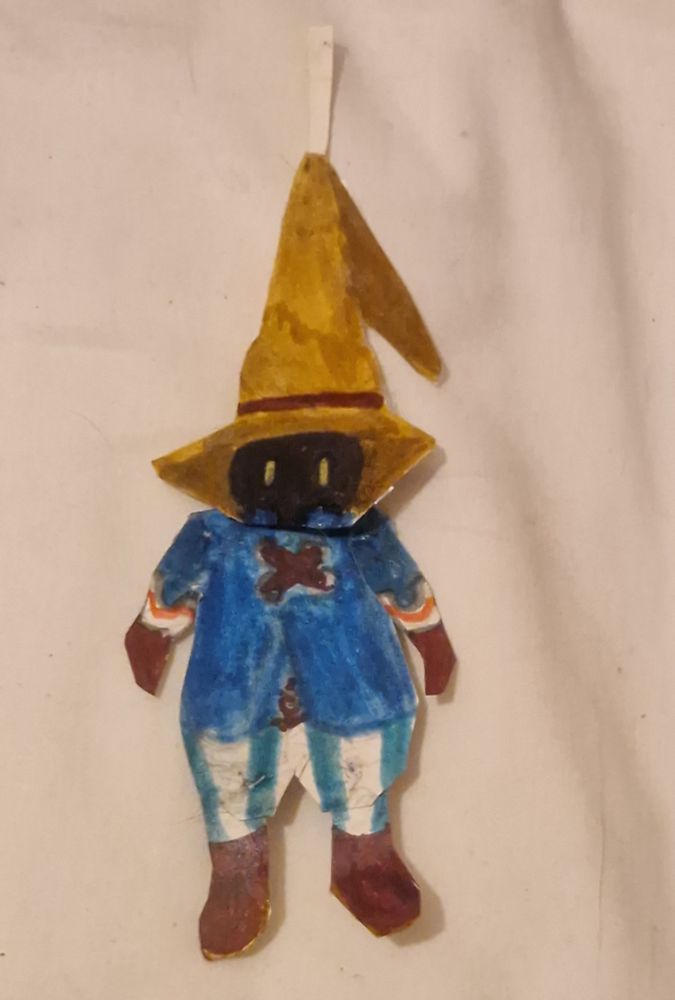 A paper doll of Vivi from FFIX. Vivi is a black humanoid creature with yellow eyes. He is a small child who wears a beige wizard's hat with a brown band, a blue jacket with brown pullstrings and white sleeves with a red trim. He is also wearing white and turquoise stripy bloomer trousers, brown gloves, and brown shoes with beige heels. 