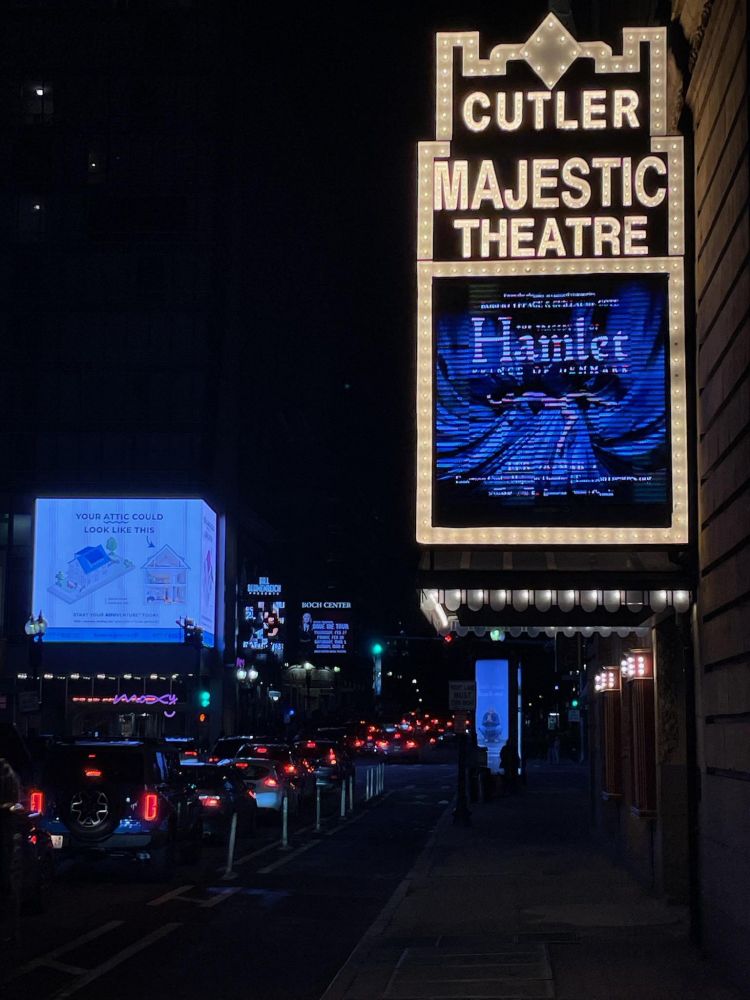 marquee of the Cutler Majestic Theater with the name of the theater in white lights and the name of the play, Hamlet, in blue lights. there is an image of Ophelia reclining. it's night, and you can see red tail lights from the cars driving by.