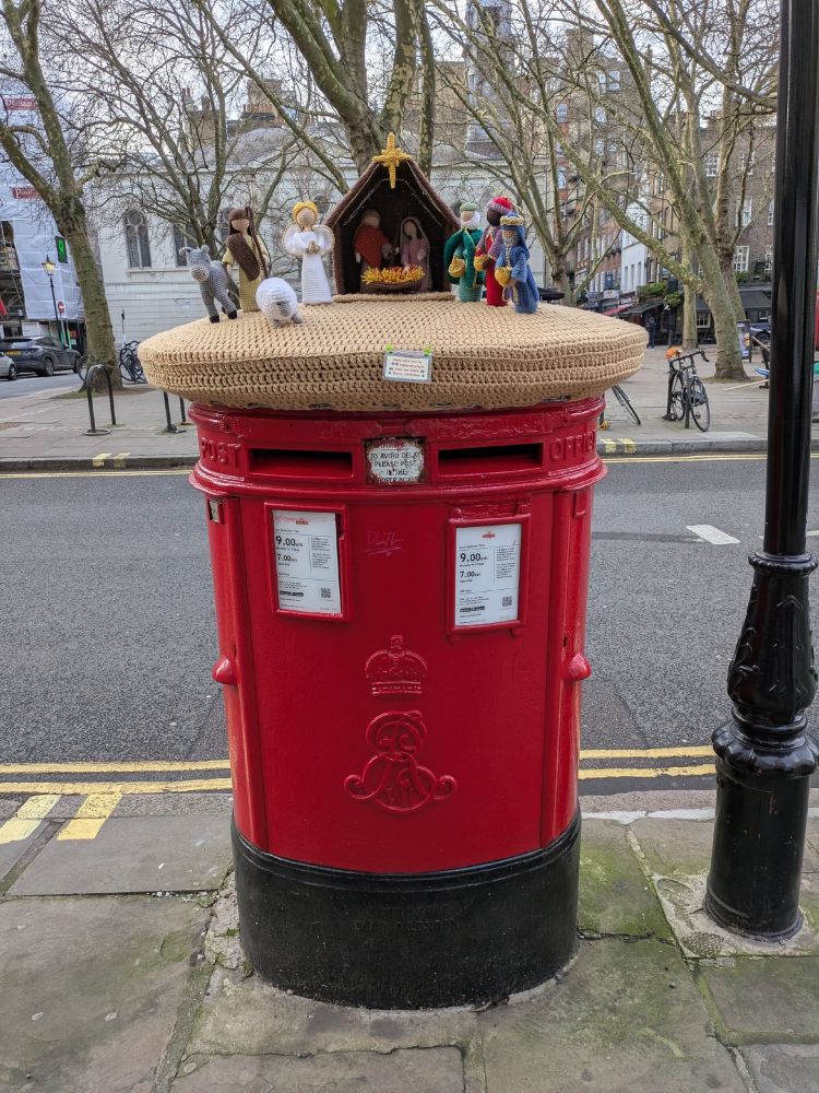 A London red mailbox with a crochet cover on top. It's of a nativity scene. 