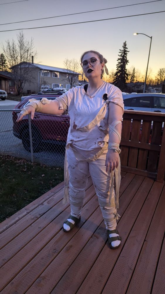 A woman stands on a wood deck dressed as a mummy.