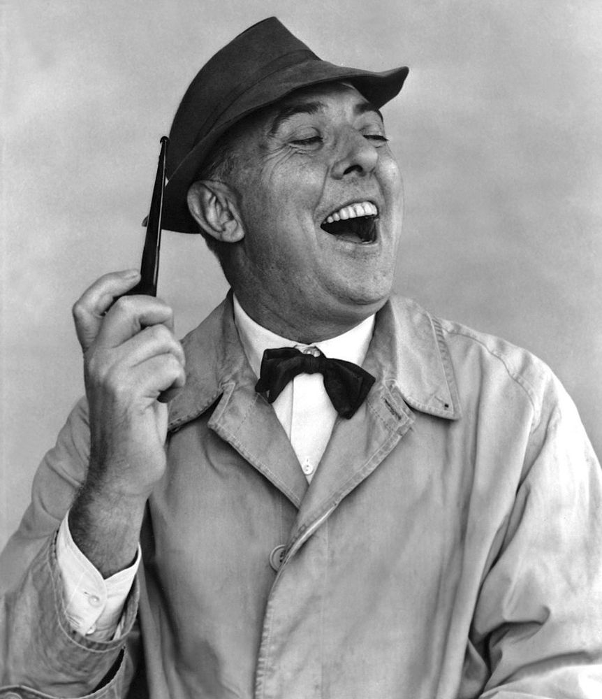 Jacques Tati, laughing, in his familiar trench coat and hat with pipe