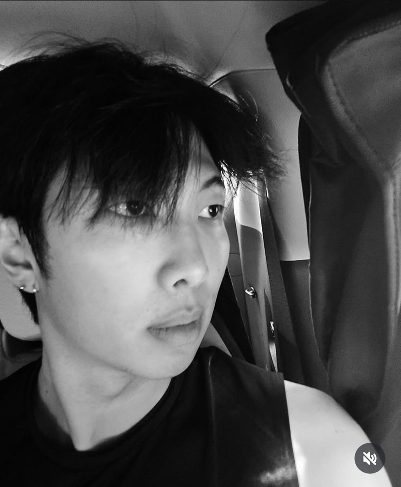 Black and white photo of Namjoon inside a vehicle, gazing out the window, looking serious with exposed shoulder. 