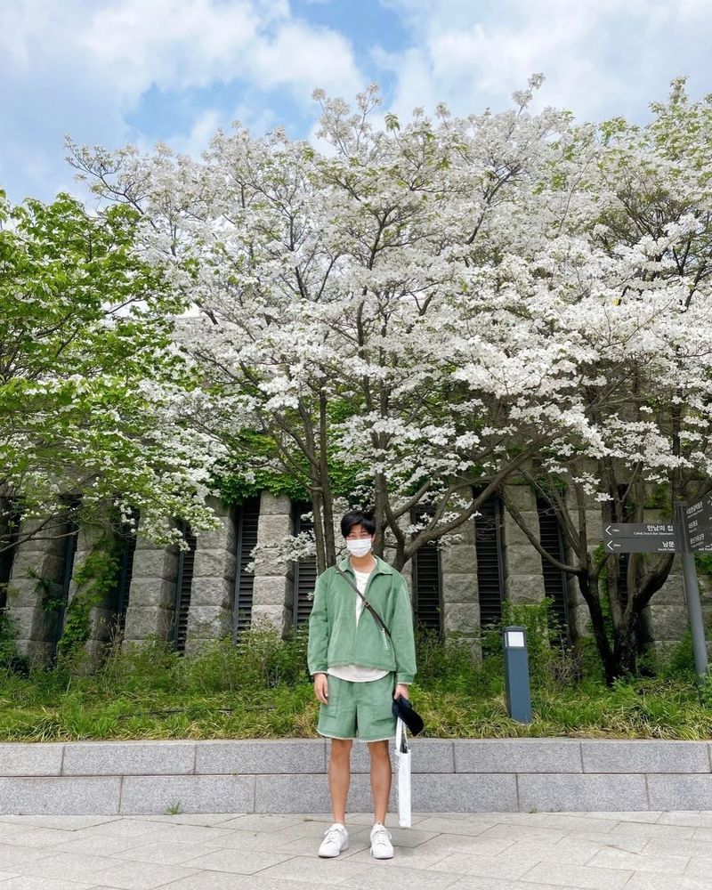 Namjoon is standing in front of cherry blossom trees. He's wearing a green shirt, green shorts, and a medical mask on his face.