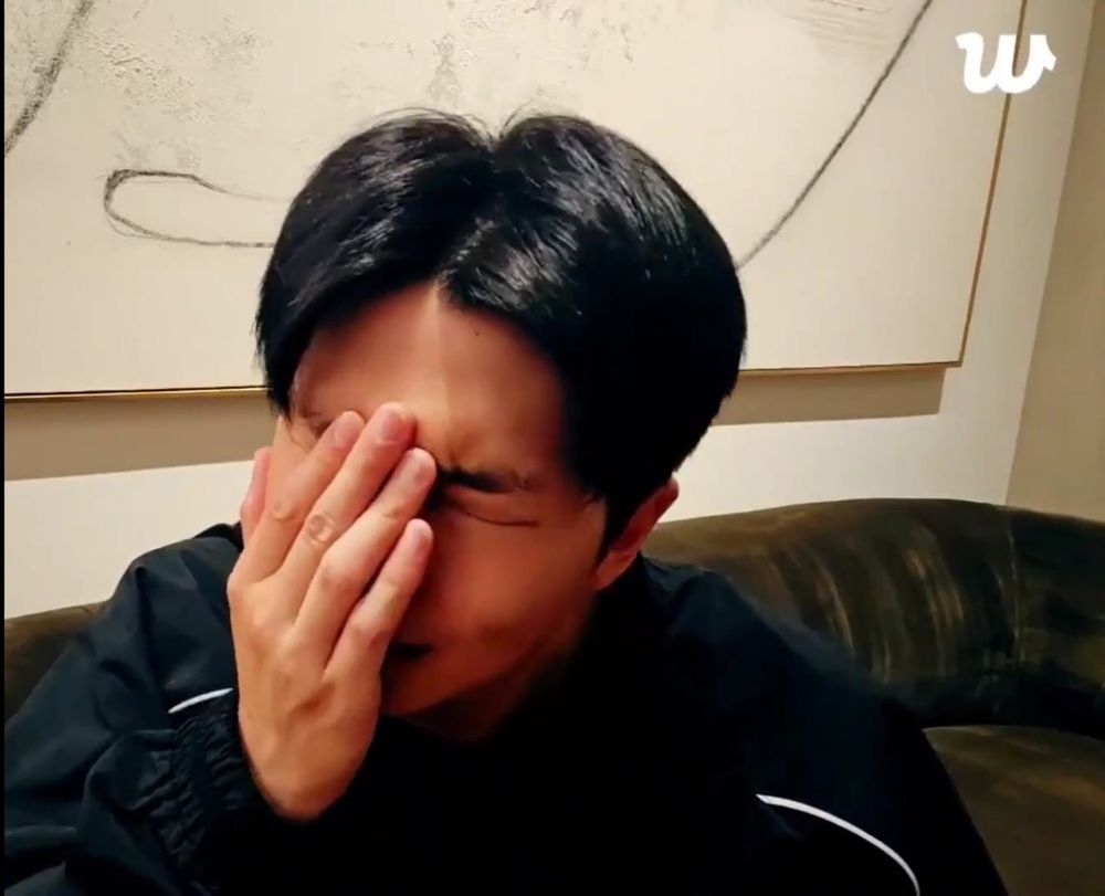 Namjoon is doing a Weverse live, sitting on a dark colored couch. There's a painting hanging on the wall behind him that seems to be white, minimalist, contemporary art. He's wearing a black jacket. His eyes are closed and his face is in his hand. He appears frustrated.