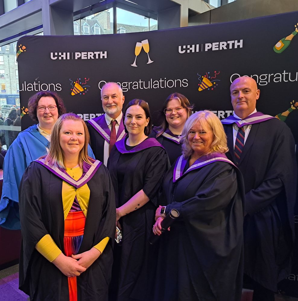 Group of people wearing graduation gowns and hoods, some are holding graduation scrolls. 