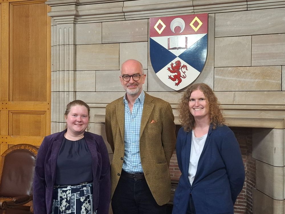 Group photo of three people standing in front of a stone fireplace, with the crest of St Andrews University above them on the wall. 