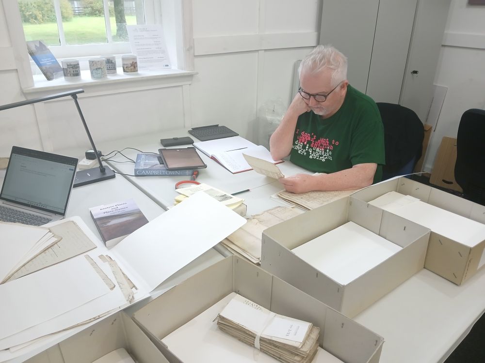 Man reading a document, sat at a table with several archival folders on it. 