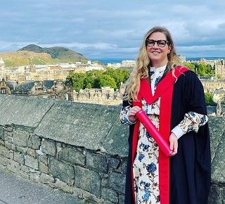 A woman wearing an academic gown with a red hood and trim, while holding a red degree scroll. Behind her is the cityscape of Edinburgh and Arthur's Seat hill. 