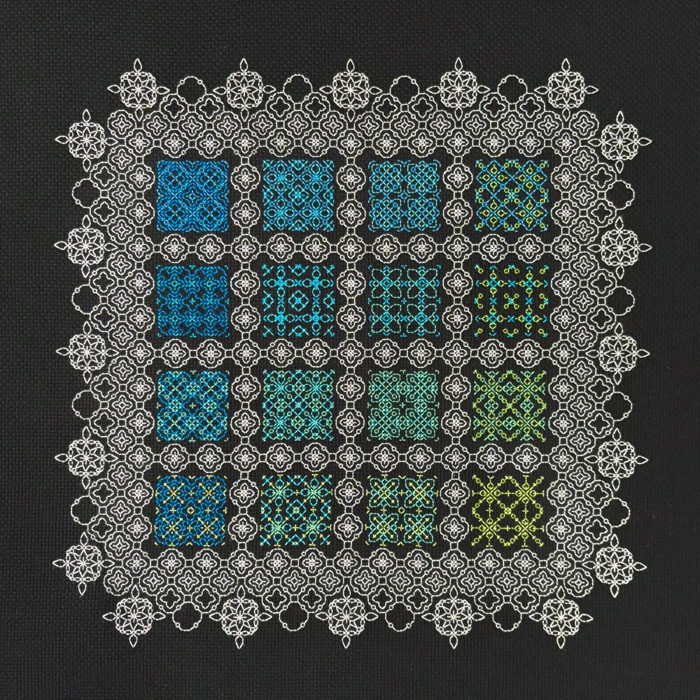 A photo of a blackwork embroidery sampler on black Aida cloth.  The sampler consists of 16 abstract geometric motifs stitched in shades of blue and green thread, surrounded by repetitive motifs stitched in white thread.