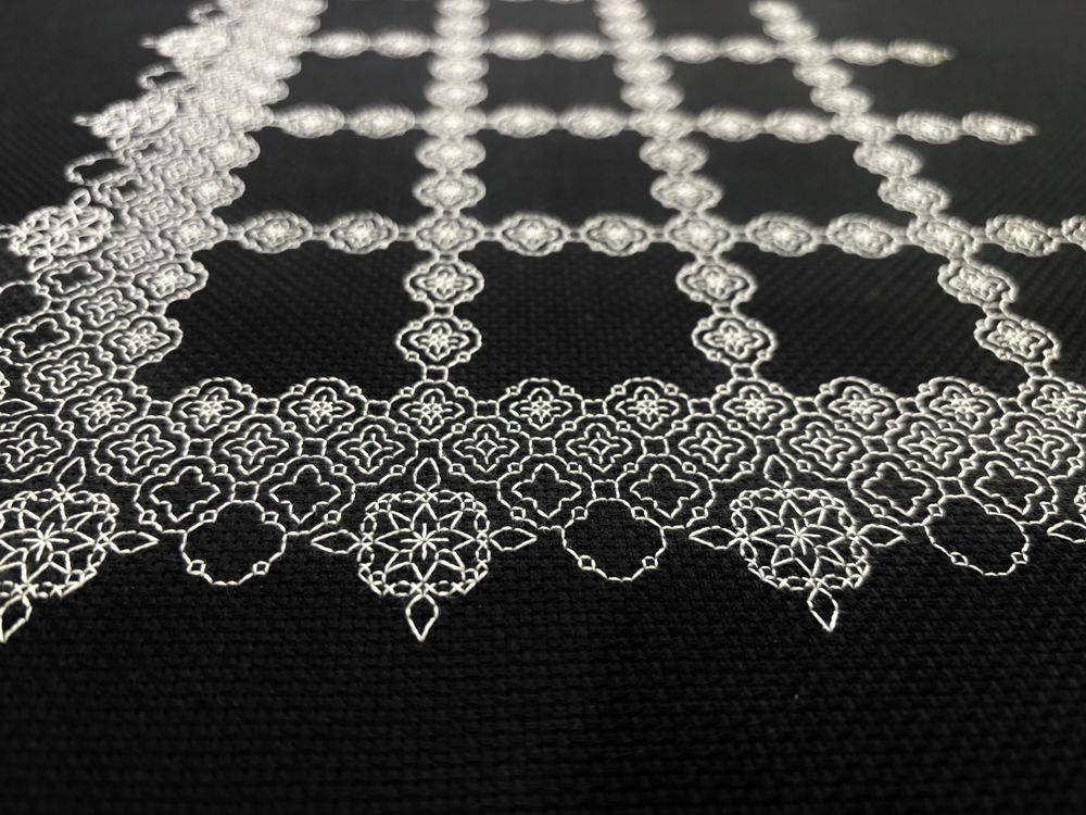 A photo of an unfinished piece of blackwork embroidery on black Aida cloth. A small geometric motif in white thread is repeated in a grid pattern surrounding blank squares that have not yet been filled.  The border consists of fading repetitions of the same motif with a larger motif at points.  The camera is held low to the cloth, close to one border.