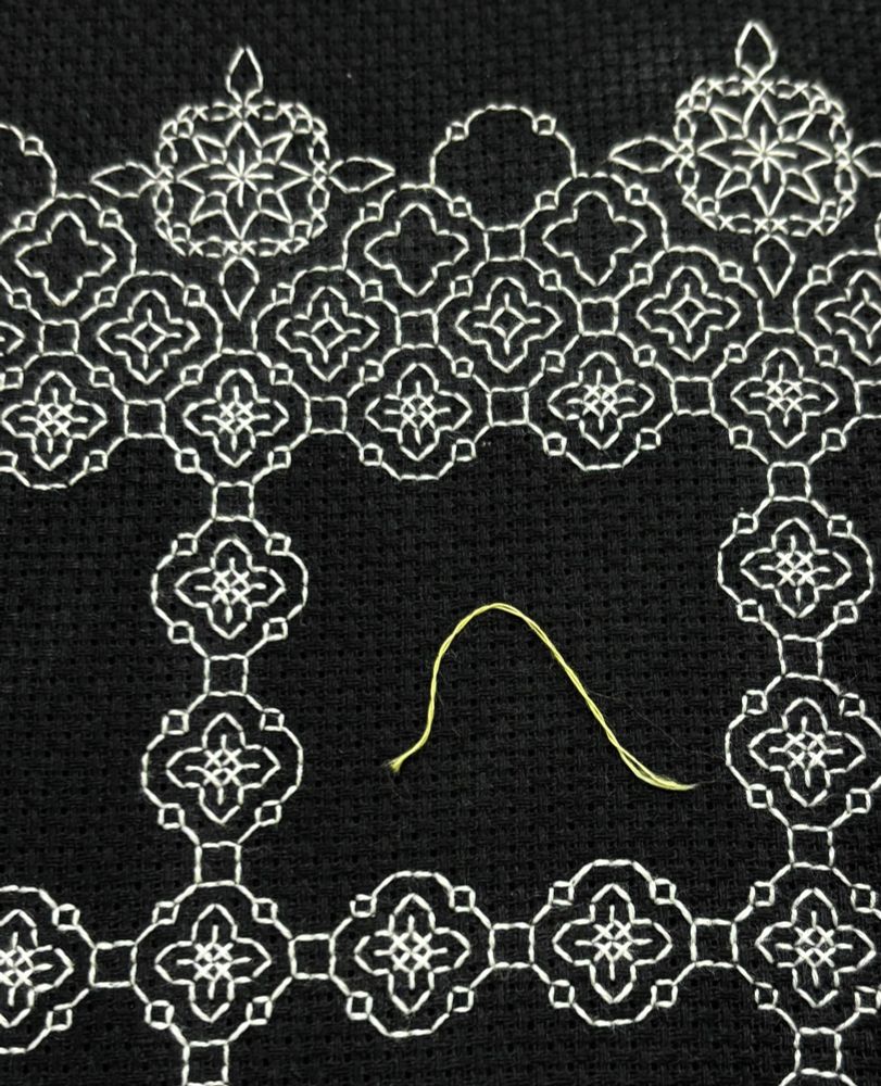 A photo of a very short piece of pale yellow embroidery floss lying on black Aida cloth.  It sits in a blank space surrounded by geometric motifs stitched in white thread.