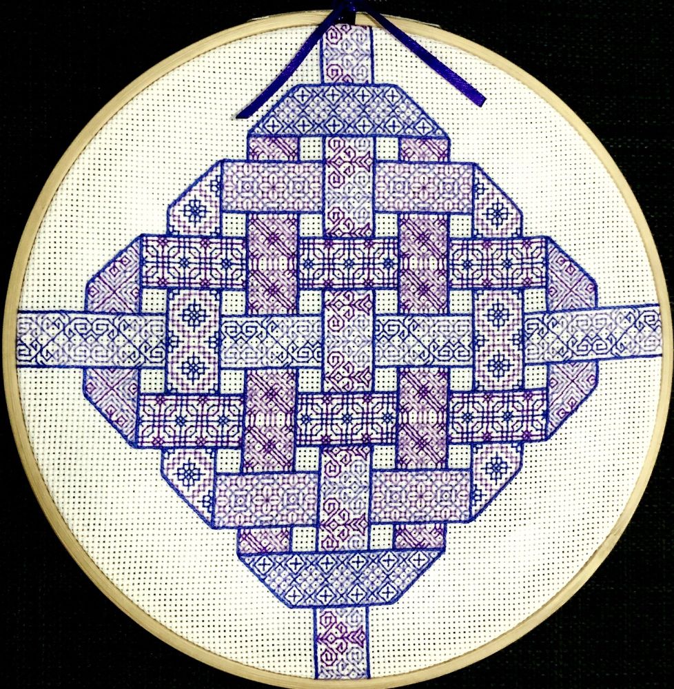 A photo of a piece of blackwork embroidery on Aida cloth.  The piece shows ribbons woven into a square-shaped knot.  The ribbons are filled with motifs in shades of purple and blue thread, and are outlined in blue thread.  The piece is displayed in a bamboo embroidery hoop and sits on a black background.