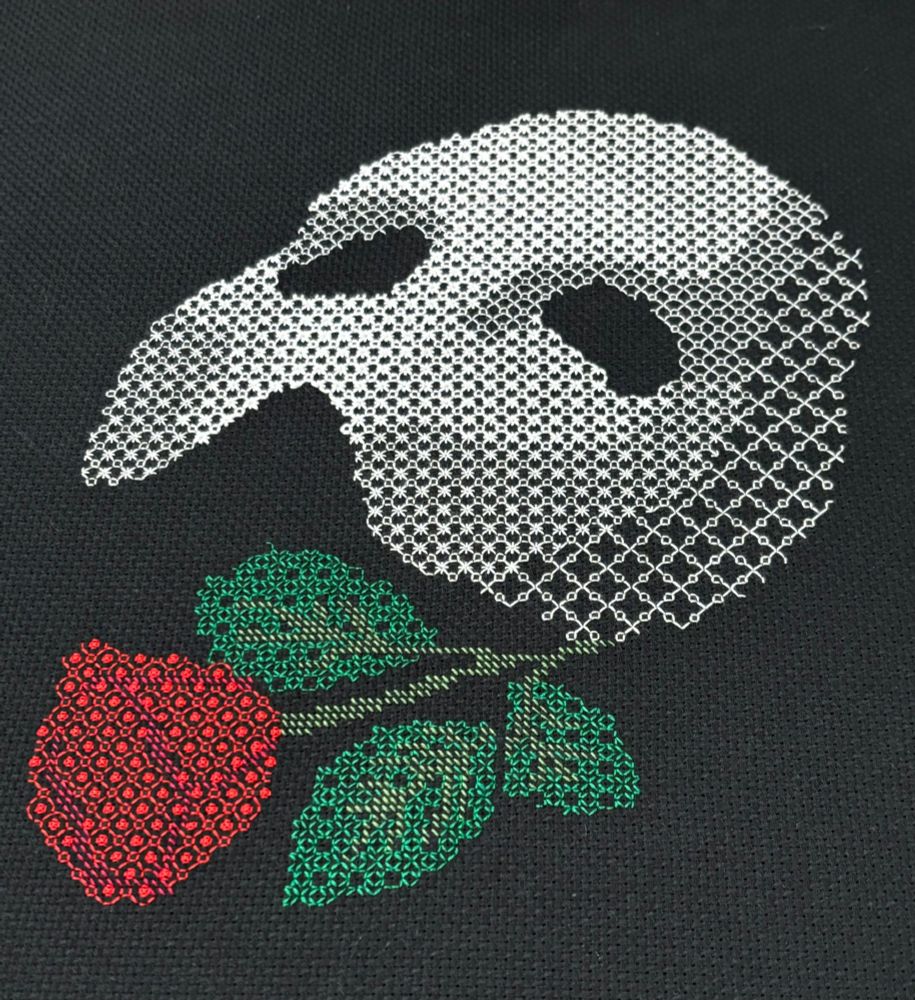 A photo of a piece of blackwork embroidery on black Aida cloth.  The piece shows the Phantom of the Opera mask (a white half-mask that would cover from the forehead to the nose) and rose.  The mask is worked in white thread; the rose and its stem and leaves are in red and green.  The camera is held closest to the rose.