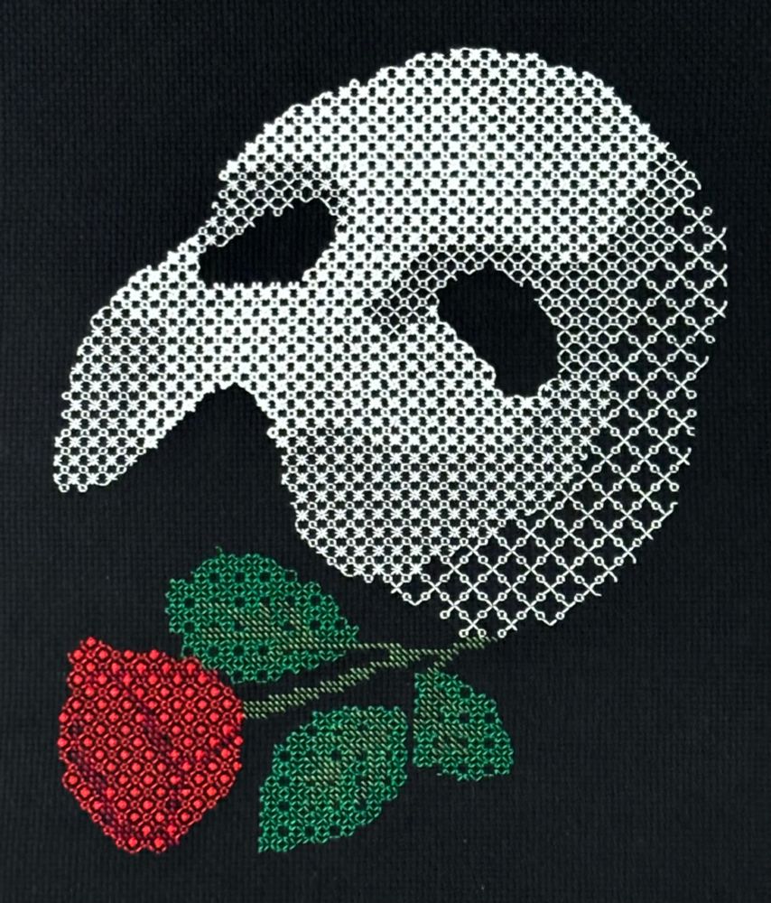 A photo of a piece of blackwork embroidery on black Aida cloth.  The piece shows the Phantom of the Opera mask (a white half-mask that would cover from the forehead to the nose) and rose.  The mask is worked in white thread; the rose and its stem and leaves are in red and green.