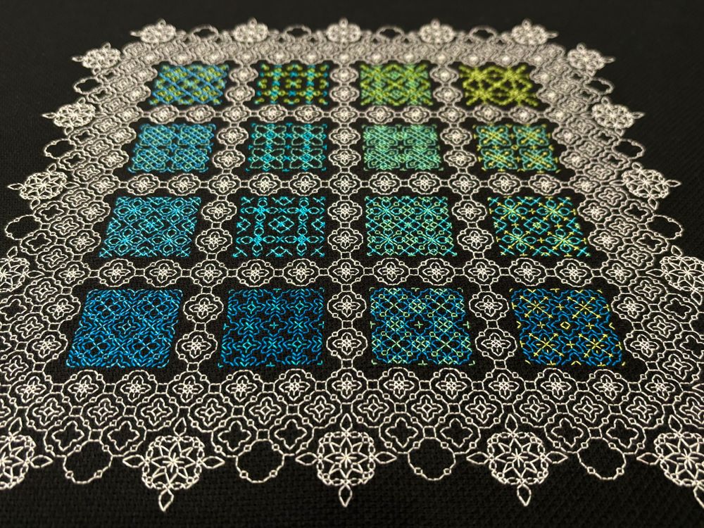A photo of a blackwork embroidery sampler on black Aida cloth.  The sampler consists of 16 abstract geometric motifs stitched in shades of blue and green thread, surrounded by repetitive motifs stitched in white thread.  The camera is held close to a row of motifs showing that there are two thread colours used in each motif.