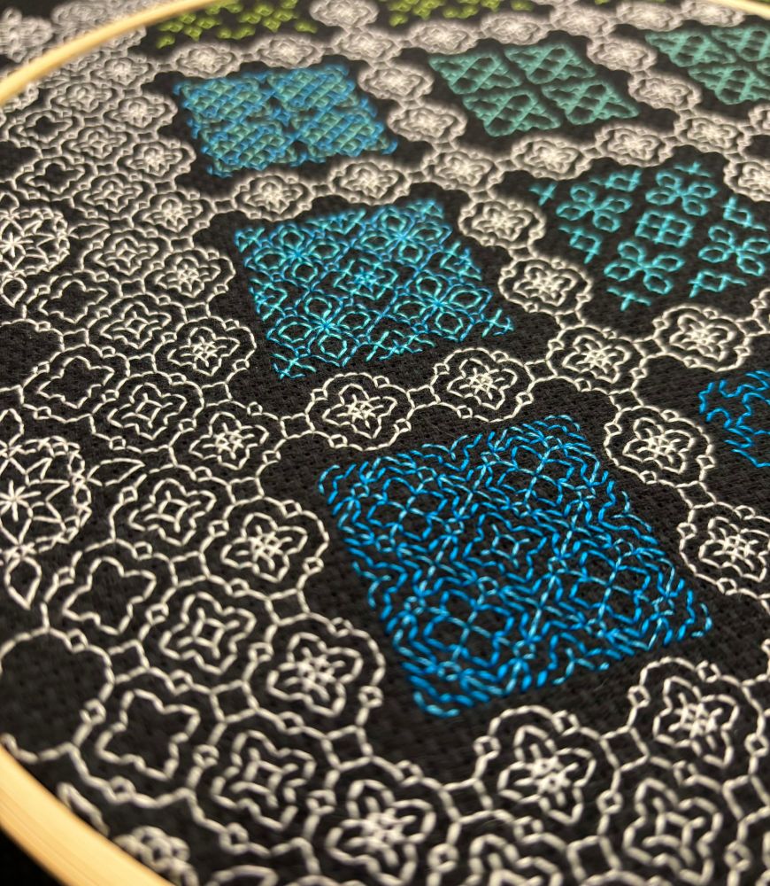 A close-up photo of an unfinished blackwork embroidery sampler on black Aida cloth.  Small geometric motifs in white thread are repeated in a grid pattern around square gaps.  The gaps are filled with motifs in blue, teal, and green thread.  The camera is closest to a square filled with two shades of blue.  A bamboo embroidery hoop can be seen around the edges of the photo.