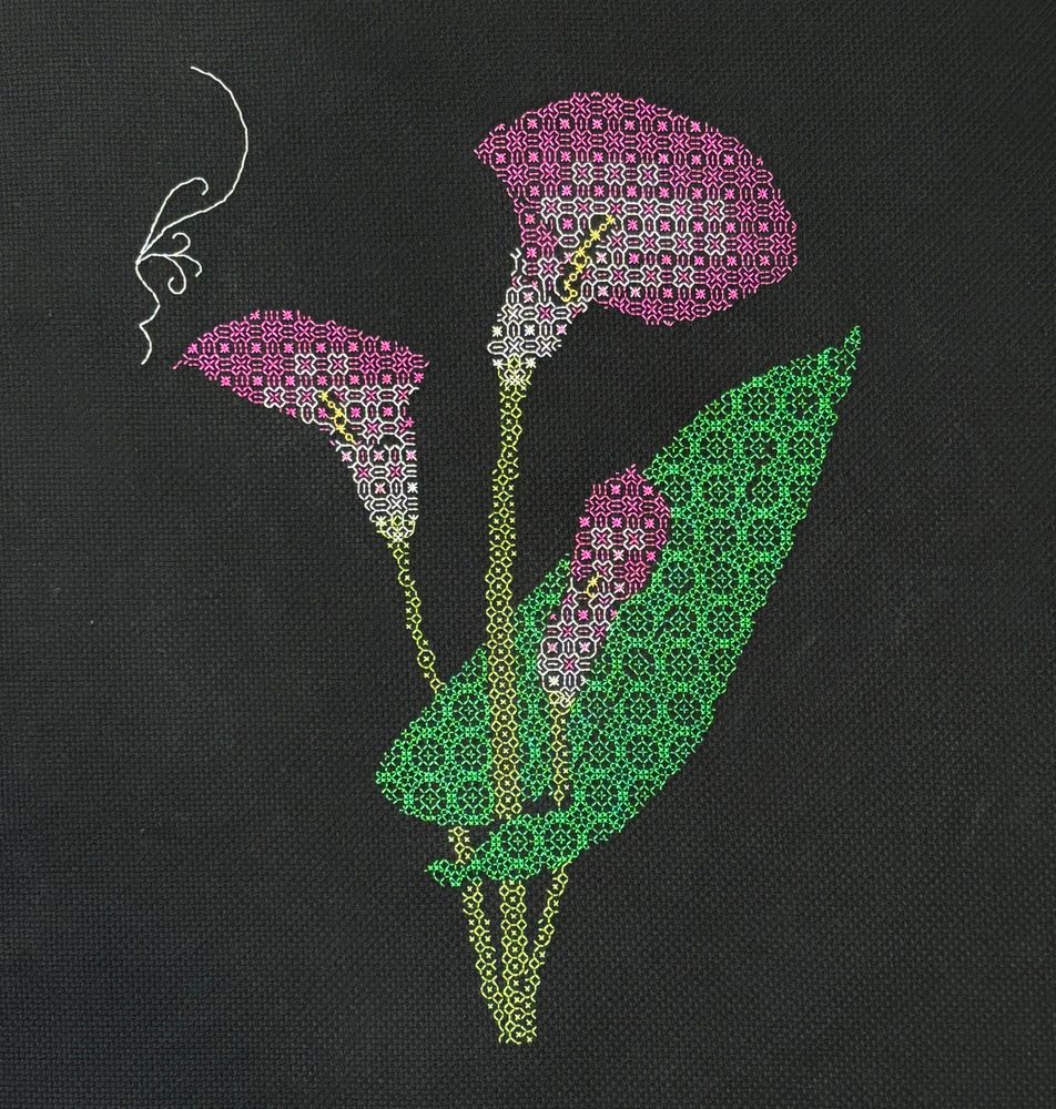 A photo of an incomplete piece of blackwork embroidery on black Aida cloth.  The piece shows three calla lilies worked in white thread at the base of the flowers, transitioning to purple thread at the outside.  The stems are shown in light green thread and a single leaf in darker green.  The beginning of a border flourish has been stitched in white thread, but is not complete.