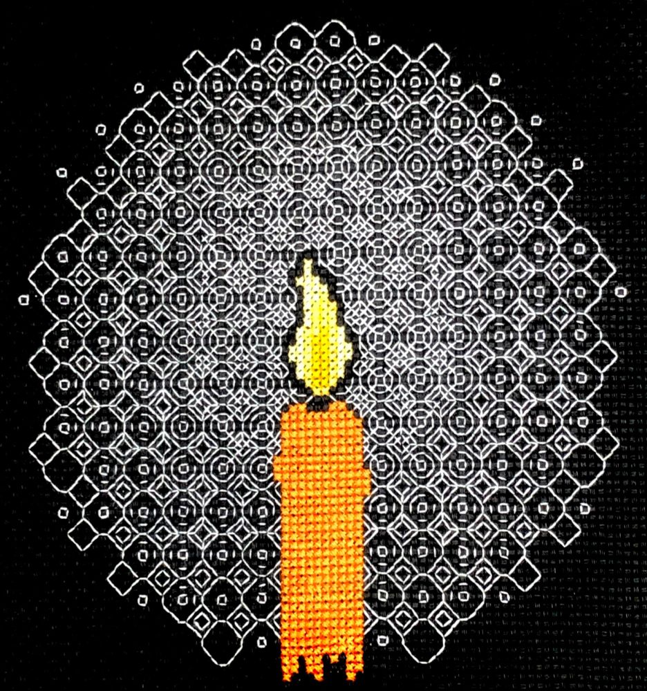 A photo of a piece of embroidery on black Aida cloth.  A candle is cross-stitched in orange thread, with the flame cross-stitched in shades of yellow.  Around the flame, a blackwork motif in white thread is repeated, fading out towards the edges of the image as if it were the light from the flame.
