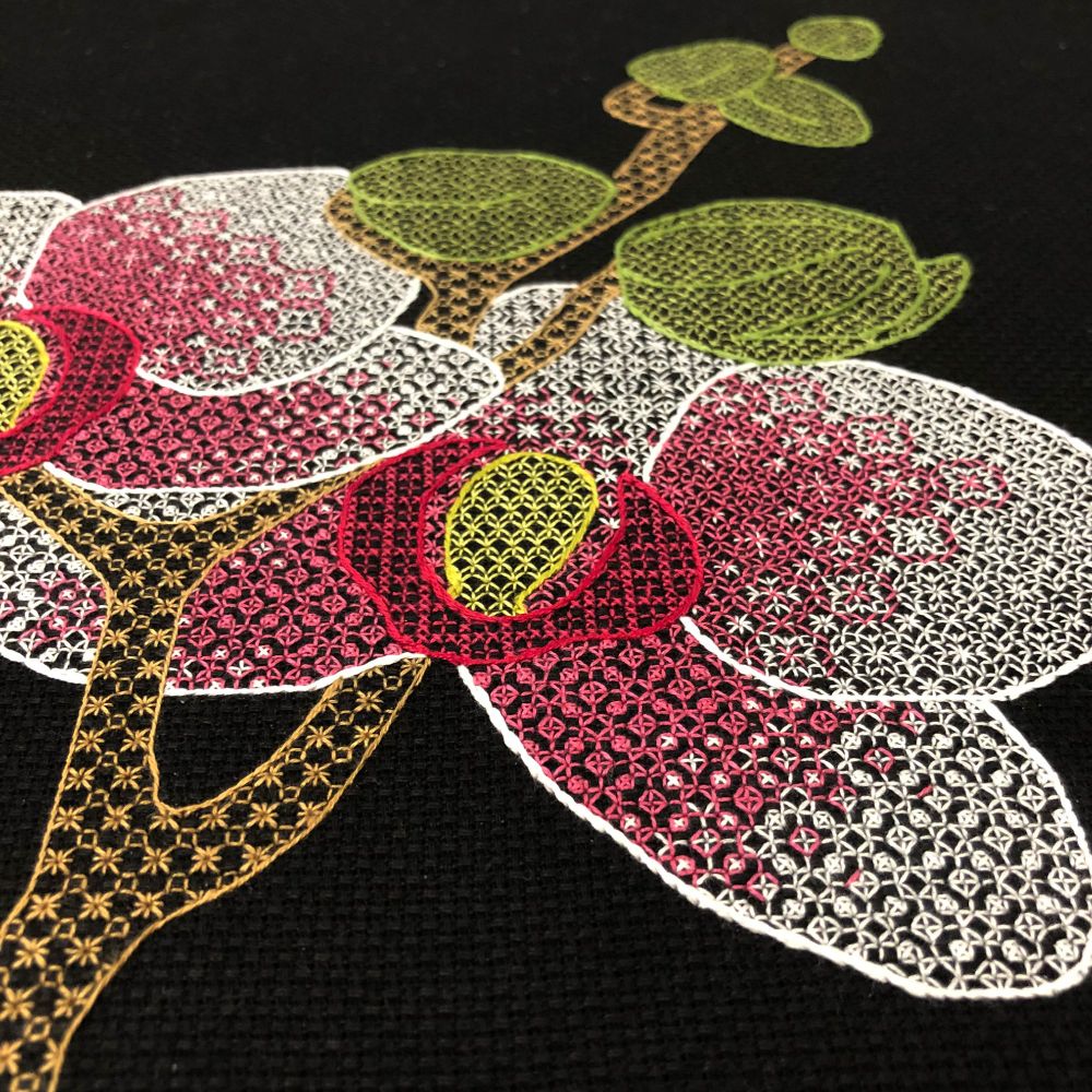 A close-up photo of a piece of blackwork embroidery on black Aida cloth.  The piece shows a stem holding two orchid blossoms with pink centres, fading to white at the outside of the petals.  Several unopened buds are farther up the stem.