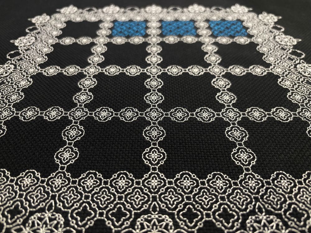 A close-up photo of an unfinished piece of blackwork embroidery on black Aida cloth.  Small white geometric motifs are repeated in a grid pattern around 16 blank squares.  Three of the squares in the most distant row are filled with blue thread, but the motif is out of focus.
