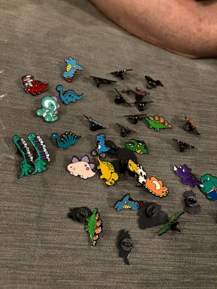 A collection of colorful cartoon dinosaur enamel pins on a gray background. Some of the dinosaurs are carrying books or have books for spikes or other body parts. 