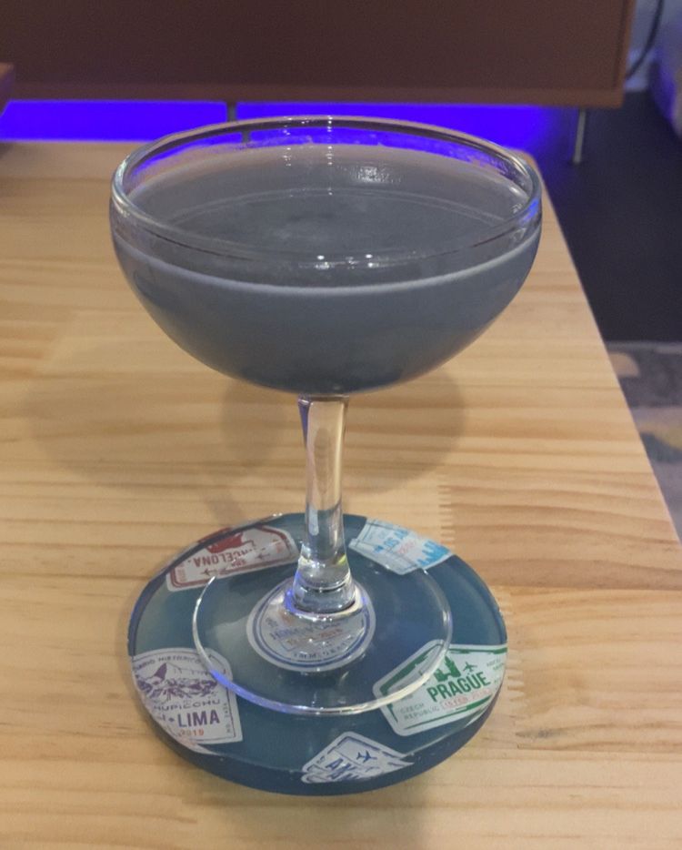 A coupe with a periwinkle colored aviation on a coaster on a light rubber wood coffee table with a darker wood colored console behind and a purple light underneath. 