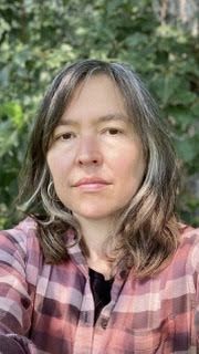 Head-and-shoulders color photo of author EC Dorgan, a woman with dark eyes and dark hair worn at shoulder length and parted on the side. She doesn’t have bangs. She wears a red-and-pink plaid over a black crewneck and stands before a backdrop of green foliage and weathered wood. She looks directly into the camera, smiling slightly.