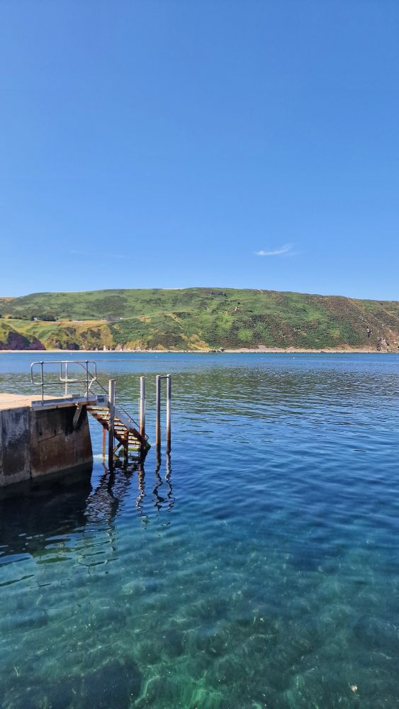 A coastal photo portrait-style with a ladder leading from a harbour wall to the left, a rocky cliff in the background and the turquoise and blue waters of the sea in the fore and midground.
