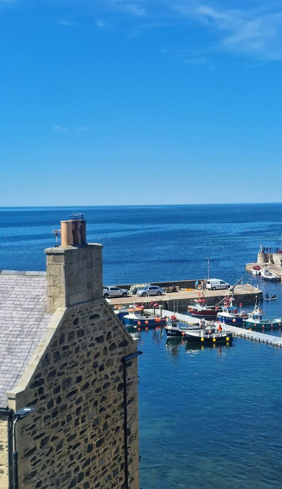 A coastal photo portrait-style overlooking a harbour from on high on a sunny day. There is the side of a stone-built house with a chimney to the left and boats, a pontoon and a harbour wall to the right.