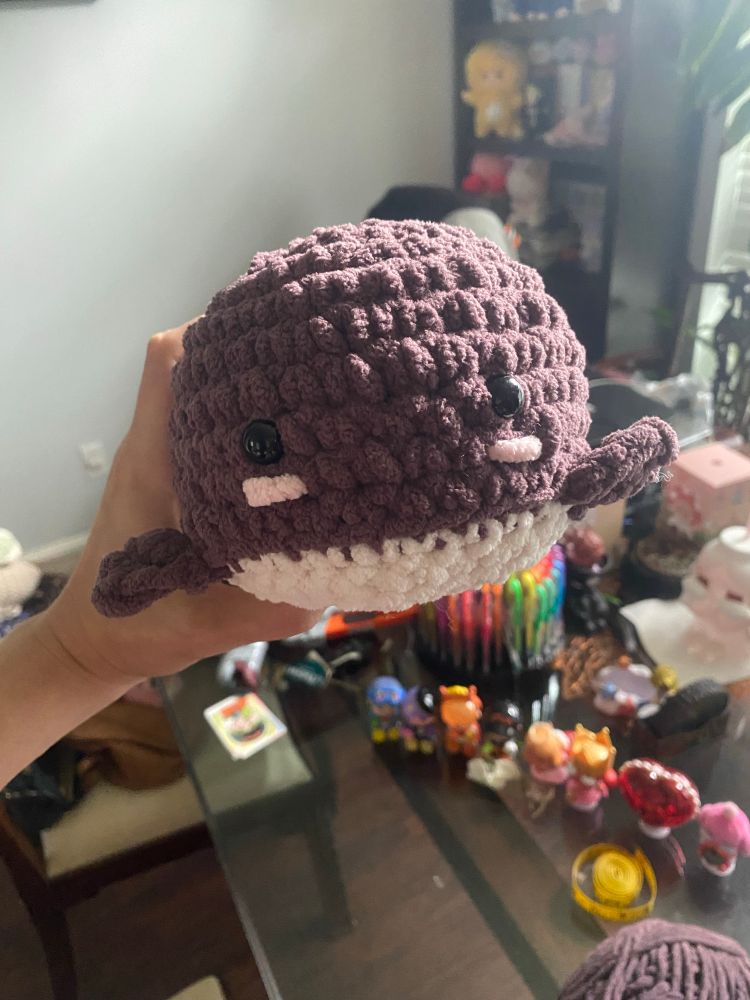 A dark purple crocheted whale with a white tummy and lil blush