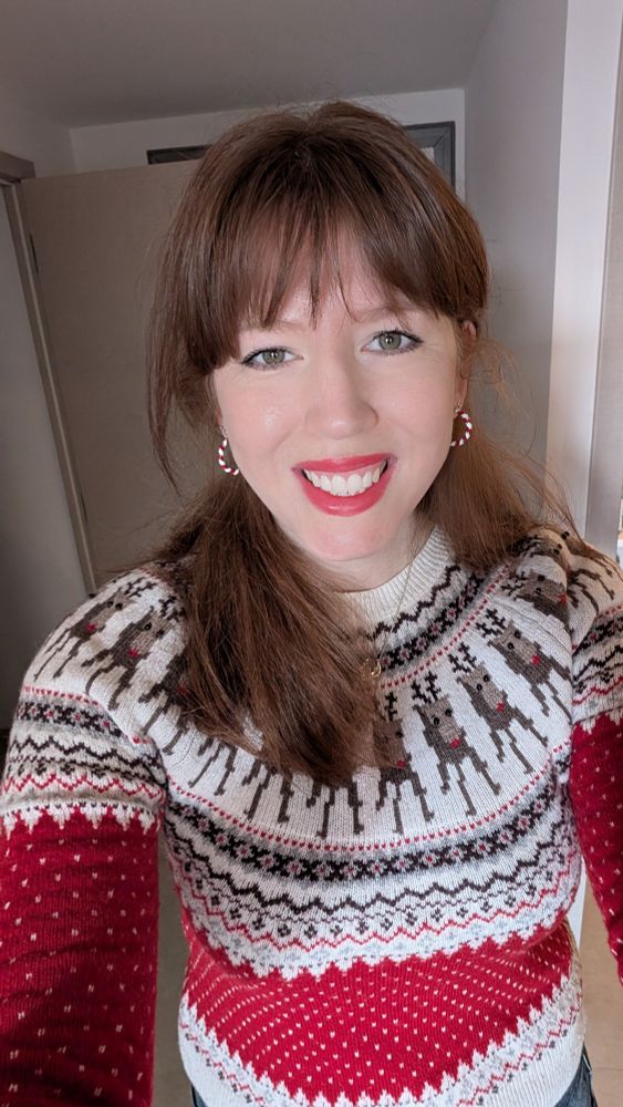 Selfie of me in a Christmas jumper with Rudolphs on it. I have candy cane earrings and red lipstick. I'm smiling at the camera. 