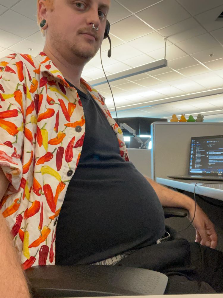 a side shot of me sitting in my work chair, my belly is in silhouette in a black undershirt with a chili pepper patterned button up over it