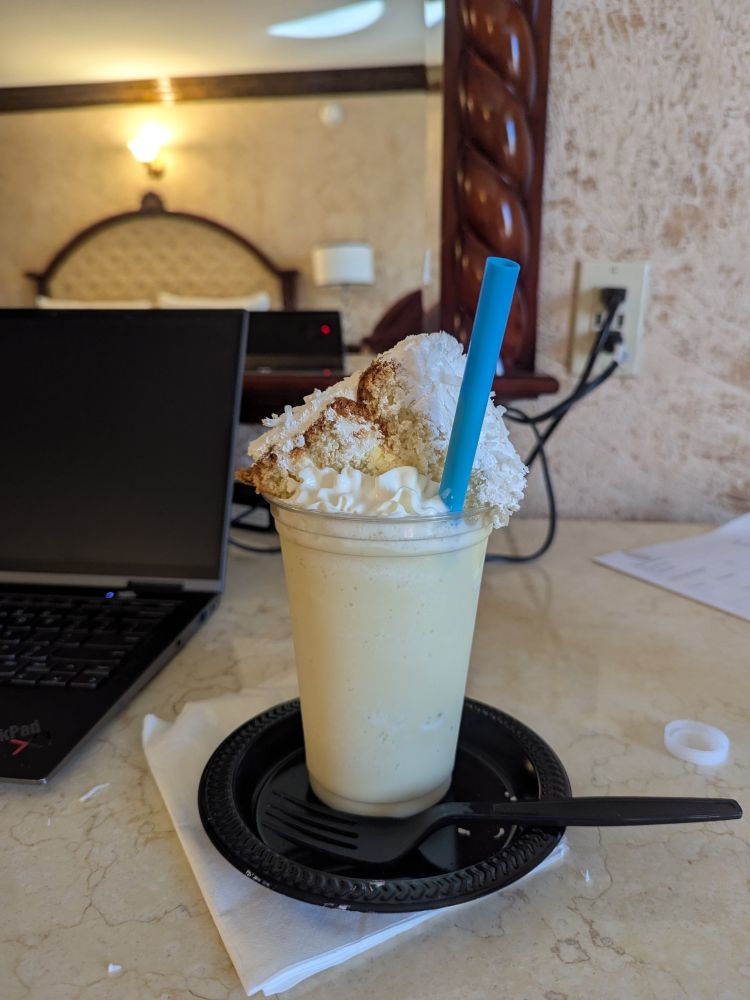 A cup full of a cream colored shake, topped with whipped cream and a slice of coconut cake