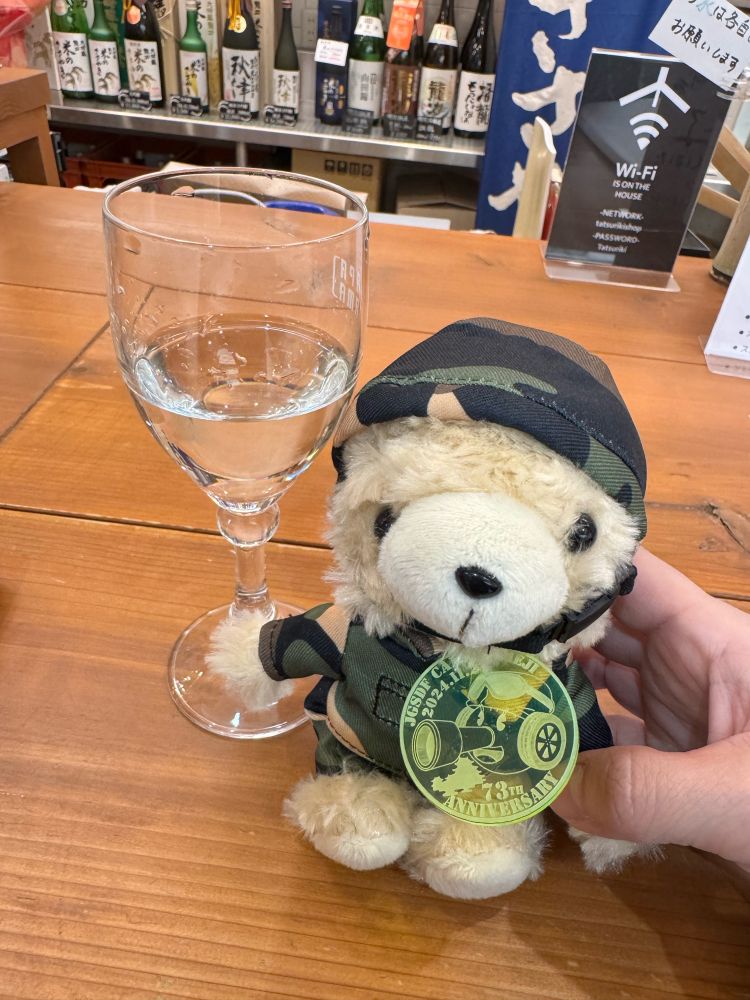 陸上自衛隊姫路駐屯地創立73周年記念行事にて販売されていた陸上自衛隊員の衣装を纏ったクマのぬいぐるみ。首元に当駐屯地のキャラクターが彫られたプレートを下げている。
隣には龍力の米のささやき ひやおろしがグラスに入っている。
撮影場所は龍力のお酒が楽しめる立ち飲み屋、玄妙庵。日本酒が好きな投稿者のお気に入りの店である。