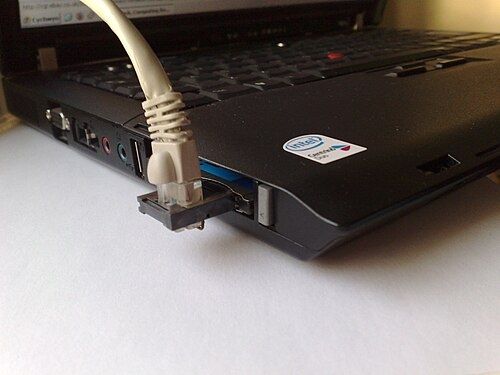 An older laptop viewed from the lower left corner. Inserted is a thin profile (single thickness) PC Card for networking. It has a network cable (ethernet) plugged into an XJACK, where the connector extends out from the thin card and the cable connects perpendicularly, poking out through the bottom of the jack uncomfortably.