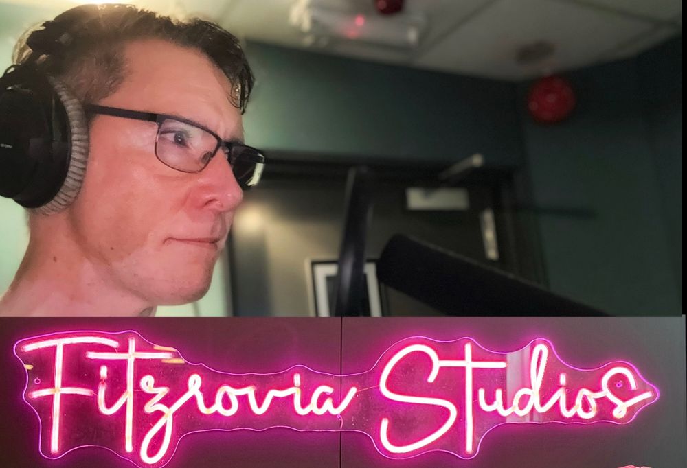 A head-shot photo of voice actor, Scott Joseph, on the left and in profile, staring with keen eyed intensity at something, probably a monitor with fabulous lines of dialogue on it, that’s off picture towards foreground right. Scott is wearing his dark rimmed glasses and his salt and pepper hair is imprisoned under a set of studio headphones. There’s a shotgun studio mic and popshield coming up from the foreground bottom right of the photo. Along the very bottom of the image Scott’s been getting all ‘arty’ by cutting in part of another photo - that of the name ‘Fitzrovia Studios’ in cursive writing as a pink neon light sign.