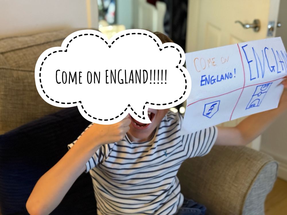 A photo of my son, whose face is obscured behind a speech bubble that reads ‘Come on England!’ And he is holding a hand made banner that he did that reads Come On England, has the three lions badge and a footy pitch drawn in it. 