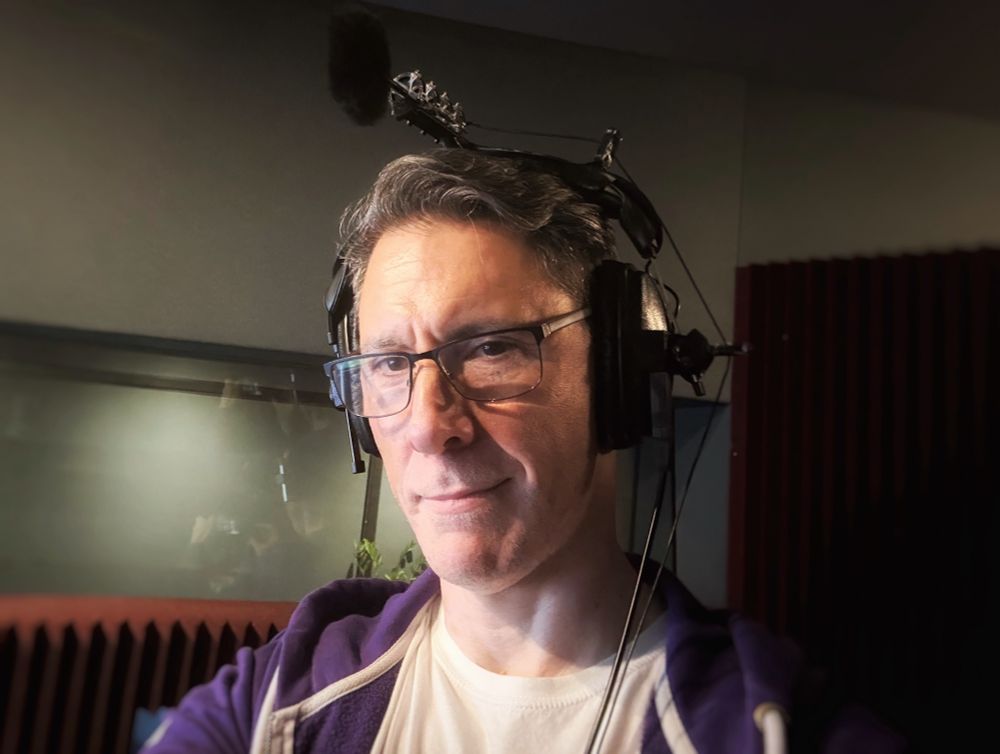 Head and shoulders shot of voice actor Scott Joseph in the centre of the photo facing foreground left. He is wearing, you got it, his white t-shirt purple hoodie combo! Seriously, he doesn’t plan it this way. Also he has on his dark framed glasses. His expression is a warm half smile trying to convince that he can actually pull off the look of this headgear (he can’t but let’s not go there). Regarding the headgear - it’s a pair of over-the-ear headphones with a microphone setup attached at the top. Scott is in a dimly lit voice recording booth with audio treatment on the walls and behind him is the main studio beyond an acoustic window.