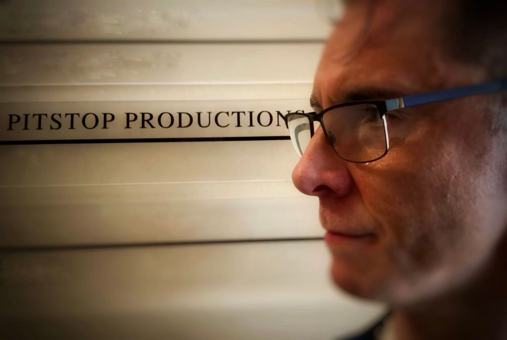 Voice Actor Scott Joseph, in an overly dramatic profile pose. His head is in the right of this photo and he is staring left with a stoic expression. All but his eyes is blurred slightly and in the left of shot is the sign in blue on off white; ‘Pitstop Productions’. It’s hard to tell from the photo but Scott is ruing the fact that he did not remember to get a more interesting photo while in the actual offices, instead he has squeezed every last drop of drama out of this photo.