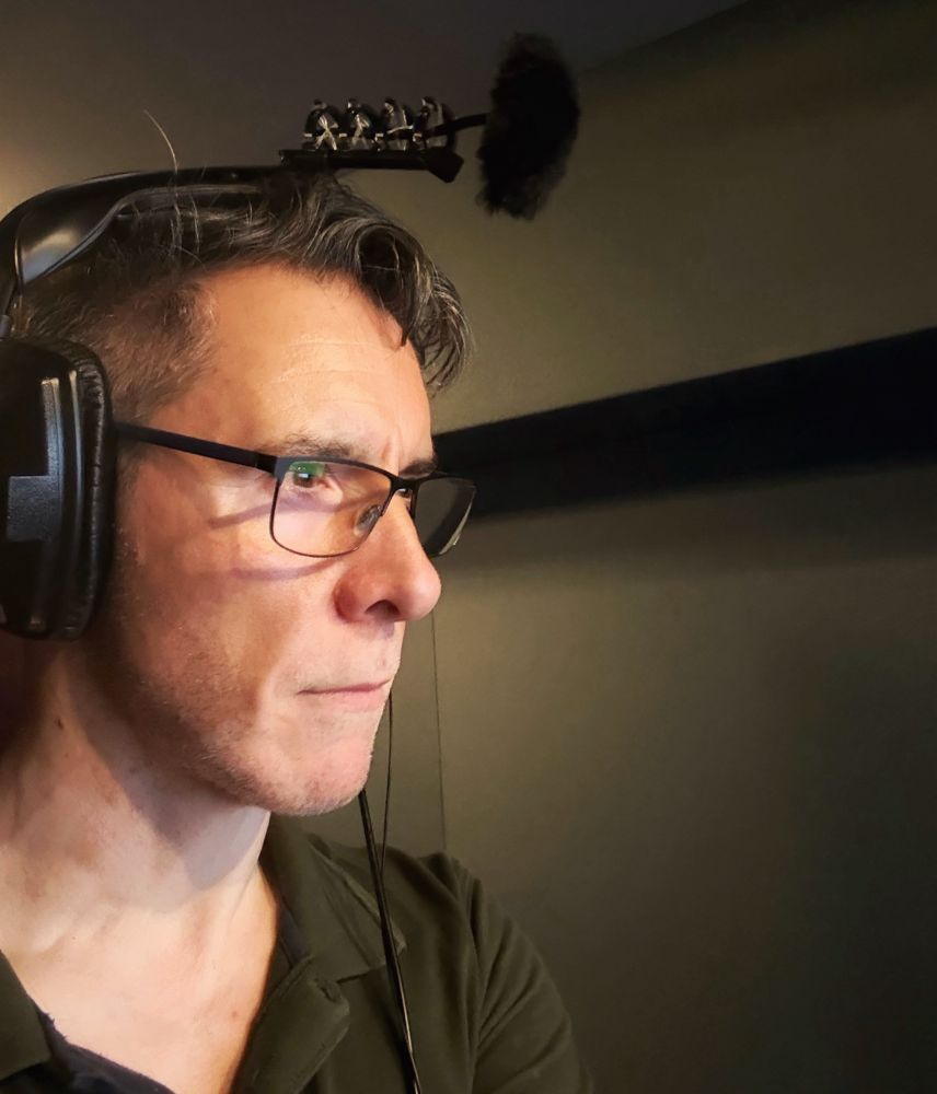Head and shoulders shot of voice actor Scott Joseph in the left of photo facing foreground right. He is in profile and wearing a green polo-t as well as his dark framed glasses. His expression once again is extremely focussed (keen eyes like those of Dunedain bowman closing in on his quarry) staring intently at something off to the right - perhaps some incredibly small text. On his head are a pair of over-the-ear headphones with a microphone setup attached at the top. Scott is in a dimly lit voice recording booth with audio treatment on the walls.