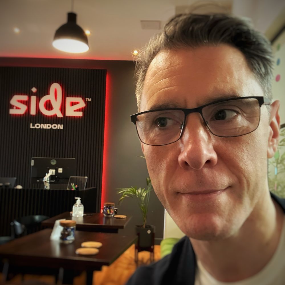 Head and shoulders shot of voice actor Scott Joseph in white t-shirt under a thin navy cardigan and he’s wearing glasses. His expression is a tight lipped smile as he is focussed on the VA session ahead but he is also mightily impressed by the brand spanking new sign Side Global has above its reception desk in the back ground. Behind him in the left of shot and beyond desks is the reception. The wall behind has a section of narrow wood strip panelling that has red lighting around the edge. On the panelling is the new logo for Side London in white lettering, the word Side also has red light glowing behind it. … and it’s almost as if Scott is looking at the sign even though it’s actually behind him, cos he’s all arty like that* 

*Scott is aware that this is not at all arty.