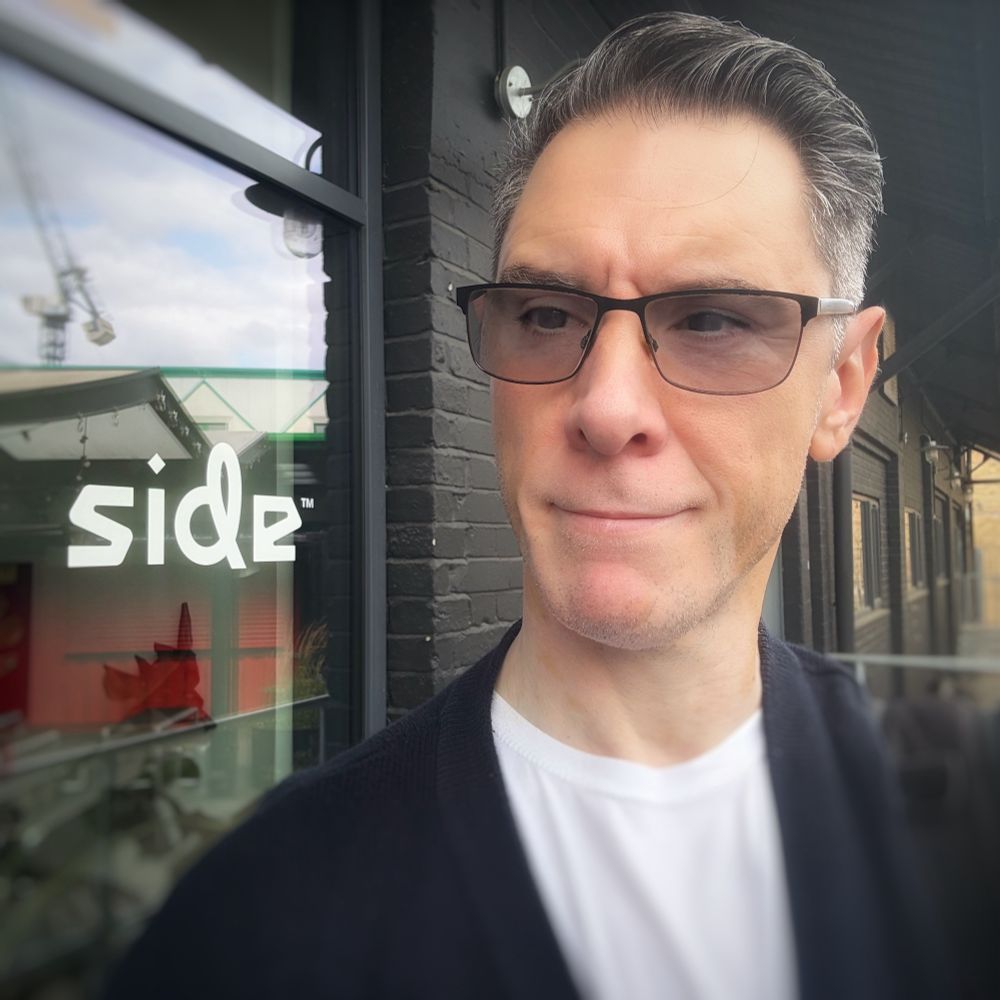 Voice actor Scott Joseph in a head and shoulder photo. He is standing right of centre, his short salt and pepper hair swept back into a side parting. He’s wearing a white T-shirt under a navy blue cardigan, together with glasses that are slightly tinted. Over his right shoulder in white stylised lettering on a glass door is the word ‘side’ - the name of the studio he’s working at. Scott is pretending to look at the sign, even though it’s behind him. However, with his expression being slightly obscured behind his tinted glasses, he looks a little like he’s crossed eyed and staring at his nose with a tight lipped smile … clearly a face for voice work! 