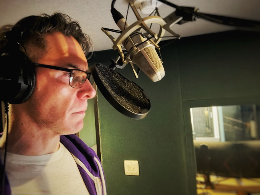 A professional recording booth at Pitstop Studios with acoustic treatment on the walls and ceiling. Background bottom right of the photo is an acoustic constructed window through to the main studio. 
Foreground left, standing in profile in a head and shoulder shot and facing right, is voice actor Scott Joseph. He is wearing a white T-Shirt and his purple hoodie. Scott would like people to know that he does own more clothing … he just clearly has studio favourites for reasons too dull to plumb in this descriptive passage. On his head is a pair of over-ear headphones and positions in front of of him in the top centre of the photo is a microphone on a high stand and Scott’s studious facial expression is cast in a somewhat sinister light by the glow from a monitor somewhere off the bottom right of the photo. This is of course not to suggest that Scott IS sinister himself or that the character he is voicing is so predisposed, though I suppose you may prefer to draw your own conclusions in due course.
As for Scott’s salt and pepper hair, best let’s avoid describing that for it is like a wild meadow of dew burdened grass that’s been trouble by a stiff breeze… headphone hair, he’s got headphone hair!!! 