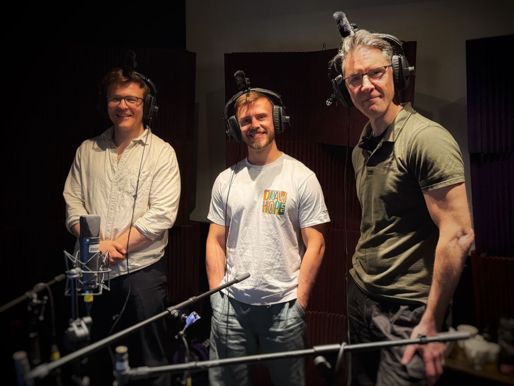 Voice Actor Scott Joseph in the right of shot posing with two other VAs Tom and Jack. They are wearing over-ear headphones with microphones on top, Scott’s is particularly prominent as it’s under a ceiling Spotlight, a detail he was unaware of when the photo was taken. 

Tom’s in the middle and wearing a white t-shirt, with his hands in his jeans pockets. Jack’s left and is taller. The both have short light brown hair. Jack’s wearing a cream linen shirt and dark trousers. Scott’s the tallest, his smile is laced with questions around why this is often the case. Three redundant mic stands are bowing before the VAs with shotgun mics turned away from them at the far end.  These three VAs have relished a voice acting session where they get to bounce off the performances of others, live! 