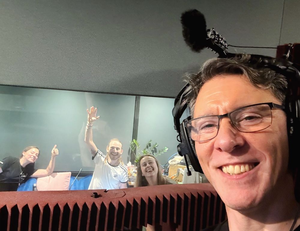 Voice actor Scott Joseph, almost looking like he’s photobombing his own photo. He is in close up popping into the picture from the bottom right and sporting a big friendly smile. On his head rather squashing his side swept salt and pepper hair, he’s wearing a pair of over-the-ear headphones with a microphone setup attached at the top. Scott is in a dimly lit voice recording booth with audio treatment on the walls and behind him is the main studio beyond an acoustic window. In the studio are three smiling faces and from left to right, they are runner Molly Tooman, director Adrian Townsend and sound engineer Polina (apologies I do not know Polina’s surname).